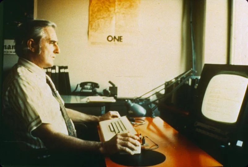 Douglas Engelbart seated at his desk at SRI, working a chord keyset beside an early CRT display